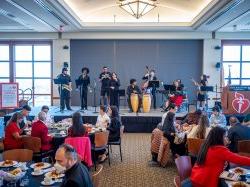 A band with bongos, brass and string instruments performs in front of a large audience at a luncheon