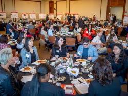 People seated at round tables eating lunch listen to a speaker during a symposium.