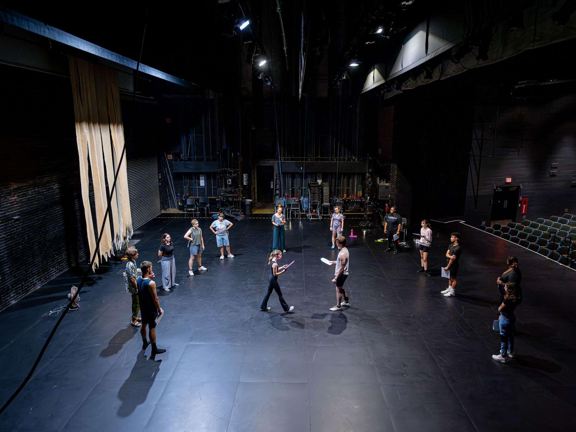 A group of 10 actors form a semi-circle on a black stage while two actors read from scripts in the middle of the stage.