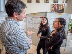 Two young scientists talk to a professor in front of a research poster.