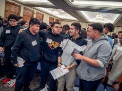 A male student put his arms around classmates in a roomful of young male high school students.