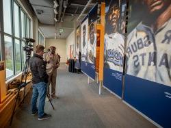 A man with a camera 和 a microphone interviews another man in front of a line of banners featuring players.